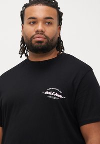 Μαύρο βαμβακερό T-shirt με κοντά μανίκια, με λευκό και ροζ λογότυπο κειμένου στην αριστερή πλευρά του στήθους. Λεία υφή και άνετη εφαρμογή.