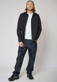 Zwarte overhemd met knopen over een wit T-shirt, gecombineerd met gestreepte blauwe jeans en zwarte sportschoenen, staande tegen een effen achtergrond.