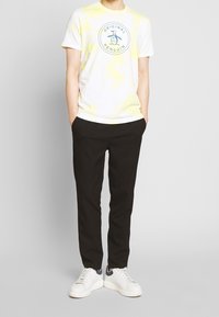 Bílá tričko s žlutým tie-dye vzorem a kruhovým logem "Original Penguin", spárované s černými kalhotami a bílými sneakers.