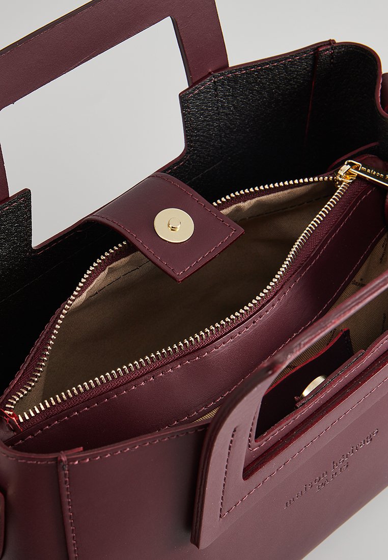 Sac à main en cuir bordeaux avec un design structuré, une poignée supérieure, une poche zippée à l'intérieur, des accessoires en or et une doublure noire contrastante.