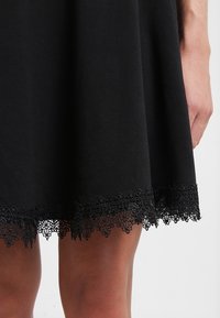 Svart kjol i mjukt tyg, med en detaljerad spetsdetalj vid fållen. Kjolen har en flödande design och sitter över knäet.