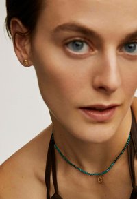 Pendientes de oro en forma de arete y un collar de cuentas multicolores que presenta cuentas turquesas y negras con un charm dorado. Piel suave, fondo neutro.