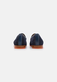 Zapatos de ante azul marino con un panel trasero de cuero suave, punta redondeada y suela de goma marrón. Diseño minimalista sin hardware visible.