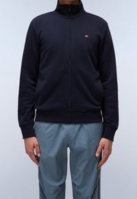 Giacca zip-up blu navy in tessuto morbido con colletto alto, polsini e orlo a coste. Presenta un piccolo logo con bandiera sul petto. Abbinata a pantaloni grigi.