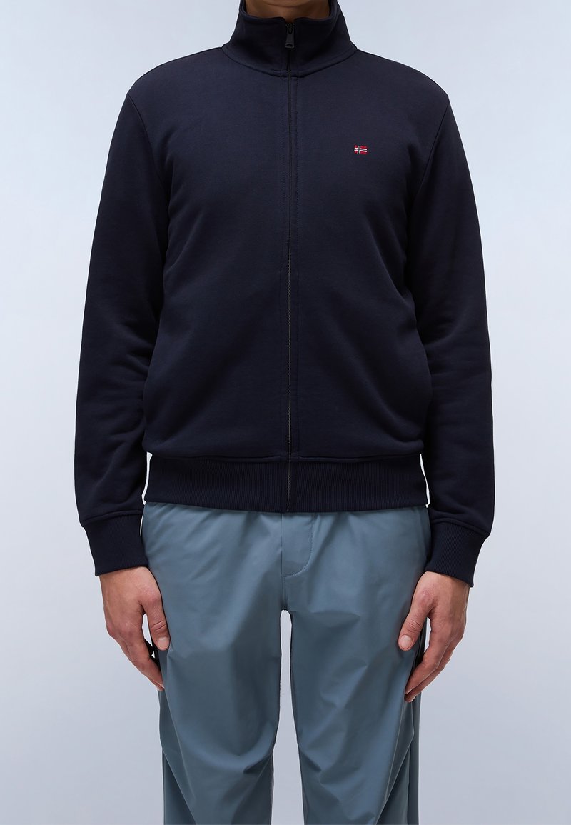 Giacca zip-up blu navy in tessuto morbido con colletto alto, polsini e orlo a coste. Presenta un piccolo logo con bandiera sul petto. Abbinata a pantaloni grigi.