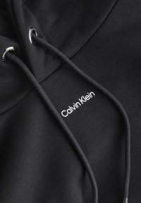 Primo piano di un tessuto di felpa nera con cordini neri e piccolo logo bianco di Calvin Klein nella zona del petto.