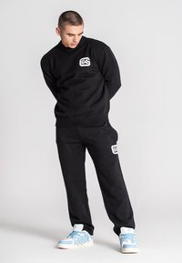 Junger Mann in schwarzem, passendem Sweatshirt und Sweatpants mit weißem Logo, schaut nach unten, trägt hellblaue und weiße Sneaker und steht vor einfarbigem Hintergrund.