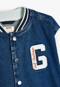 Jeansjacka med en ljusblå ribbad krage. Har en vit broderad bokstav "G" och "Golf Challenge" i orange. Snäppknäppning.
