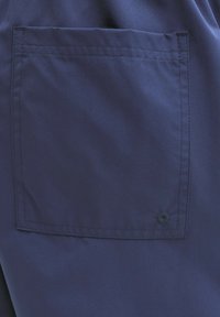 Τετράγωνη τσέπη από ναυτικό μπλε ύφασμα ραμμένη πάνω σε αντίστοιχο ρούχο με ορατές ραφές και ένα μικρό κυκλικό κουμπί κοντά στη κάτω δεξιά γωνία.