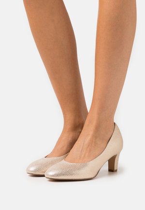 Talons en cuir verni beige avec un léger éclat, bout rond et un petit talon bloc, présentant une texture lisse et un design simple.