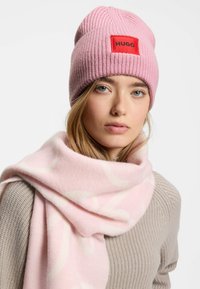 Cappello a coste rosa con toppa rossa, abbinato a una sciarpa rosa chiaro con un sottile motivo bianco, entrambi morbidi e di consistenza.
