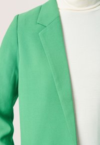 Zelený blazer z měkkého, texturovaného materiálu se strukturálním klopem, v kombinaci s krémovým rolákem. Důraz na zakázkový design a barevný kontrast.