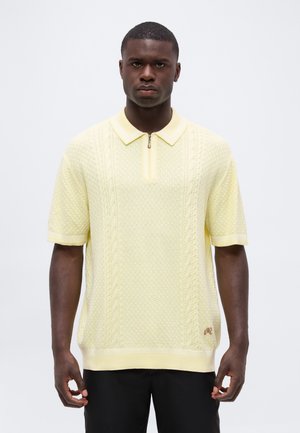Homme portant un pull tricoté jaune pâle texturé à manches courtes avec un col zippé, associé à un pantalon noir, sur un fond uni.