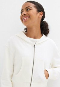 Felpa bianca con zip, cappuccio, tasche frontali e polsini a coste. Realizzata in un tessuto morbido, presenta una texture liscia e un design semplice.