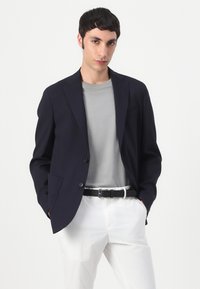 Tengerészkék blézer két gombbal, bemetszett hajtókákkal és elülső zsebekkel, szürke póló és fehér nadrág felett, fekete övvel kiegészítve.