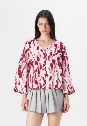 Ung kvinne med langt mørkt hår iført en oversized bluse med rosa og hvite mønstre og grå skjørt med folder, stående mot en lys bakgrunn.