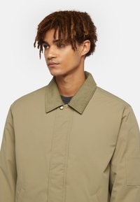 Khaki jacket with a corduroy collar, featuring a front snap closure, side pockets, and a smooth, textured fabric.