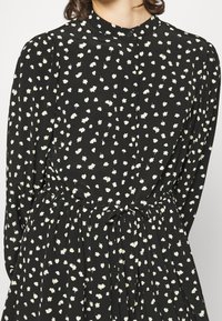 Vestido negro con un pequeño patrón floral blanco, mangas largas, escote redondo con cierre de botón y cintura fruncida con un detalle de lazo.