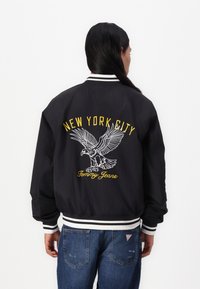Sort bomberjakke med hvid og gul broderet tekst "NEW YORK CITY" og et ørnedesign. Ribbet manchetter og kant med vandrette striber.