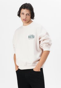 Lichtbeige sweatshirt met een relaxte fit, voorzien van een groene grafische tekst "Buena Pasta Pesto Club" op de linkerborst. Zwarte broek.