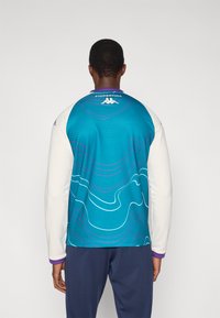 Ilgas rankovėmis sportinis marškinėlis, turintis turkio mėlynos ir baltos spalvų derinį, mėlynos ir violetinės spalvų bangų raštą, violetinės spalvos apvadais ir logotipu prie kaklo.