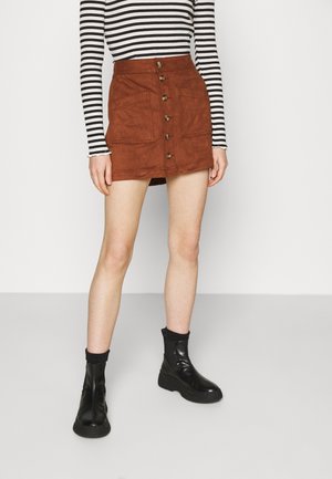 Kvinde iført brun mikrosuede mininederdel med knapper foran, sorte ankelstøvler og stribet langærmet top, stående på ensfarvet lys baggrund.