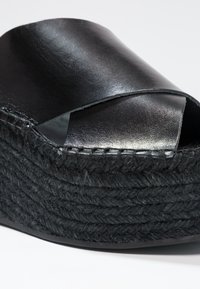 Sandale plateforme noire avec brides en cuir croisées et texture en corde tissée sur la semelle épaisse, sur fond blanc.
