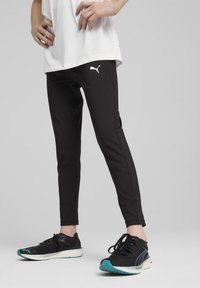 Černé sportovní legíny s úzkým střihem, se světle bílým logo Puma a kontrastními bočními akcenty, doplněné o černé běžecké boty s tyrkysovými detaily.