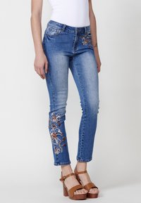 Blå denimjeans med blommig broderi på vänster ben, slim fit-design, medelhög midja och klassisk femfickestil.