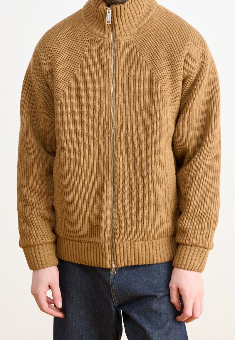 Brun ribbad stickad zip-up tröja med hög krage, med rak passform och elastiska ärmslut och nederkant, ihop med mörka jeansbyxor.
