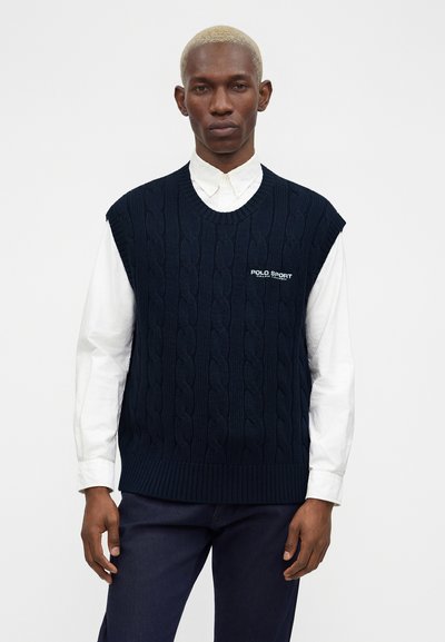 Άνδρας που φοράει μπλε σκούφο πλεκτό γιλέκο χωρίς μανίκια πάνω από ένα λευκό πουκάμισο με κουμπιά και λογότυπο "Polo Sport" στο στήθος, σε απλό φόντο.