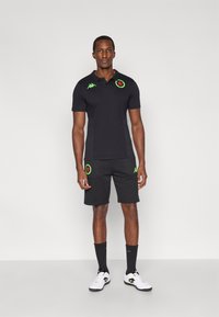 Fekete rövid ujjú póló zöld logó részletekkel és hozzáillő fekete rövidnadrág, amelyen kör alakú zöld foltok találhatók. Fehér tornacipővel és fekete zoknival viselve.