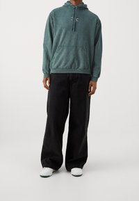 Sudadera desgastada en color teal con un bolsillo frontal y capucha con cordón, combinada con jeans negros de corte holgado y zapatillas blancas.