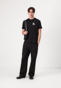 Black cotton t-shirt with white Calvin Klein logo, paired with black trousers and black shoes. The model carries a black shoulder bag.