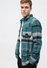 Modrá a šedá kockovaná košile z kartáčované látky, s černou náprsní kapsou, zapínáním na knoflíky a dlouhými rukávy.