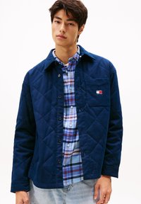 Jaunas vyras, vilkintis mėlyn�ą siuvinėtą striukę ant šviesiai mėlyno languoto marškinėlio ir šviesių džinsų, stovi prieš baltą foną.