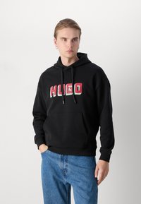 Juodas gobtuvas su priekine kengūros kišenėle, ant kurios matomas ryškus "HUGO" logotipas raudona ir žalia spalvomis. Derinamas su mėlynomis džinsomis.