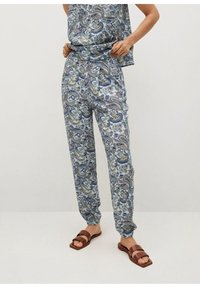 Vrouw in blauwe broeken met paisleypatroon en bijpassende mouwloze top, staat blootsvoets op een effen achtergrond.