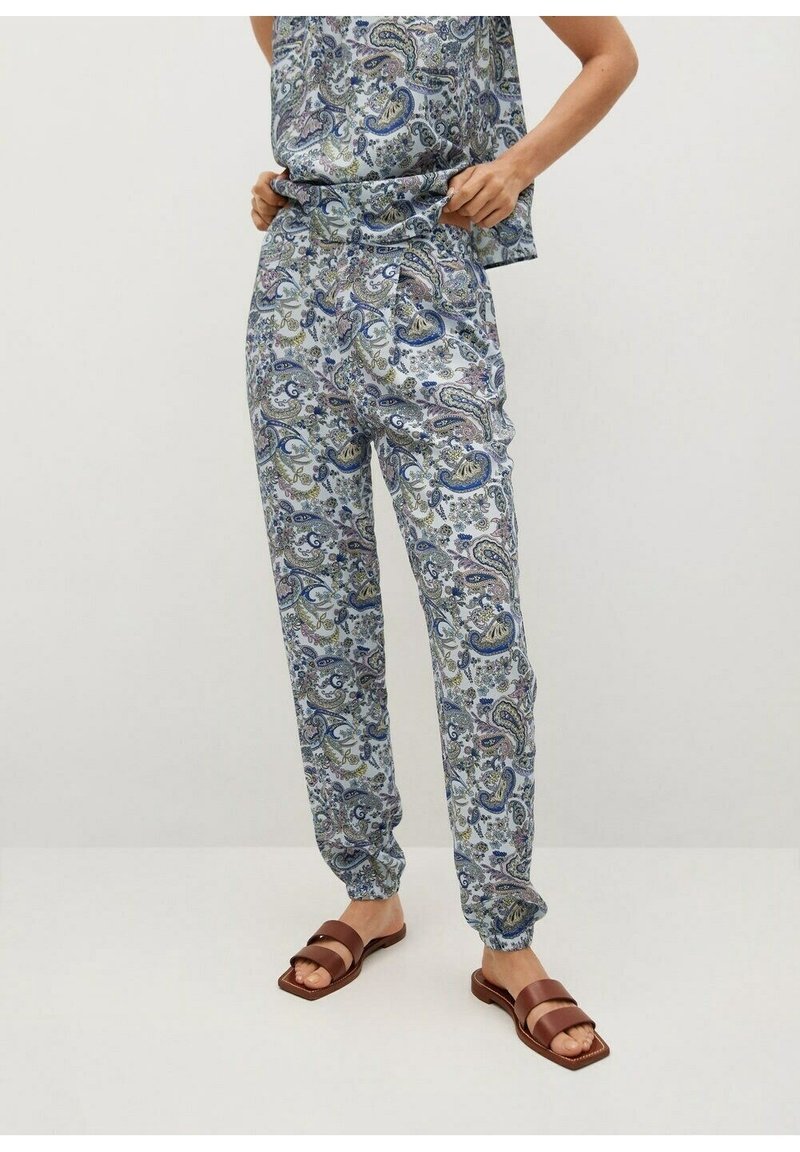 Vrouw in blauwe broeken met paisleypatroon en bijpassende mouwloze top, staat blootsvoets op een effen achtergrond.