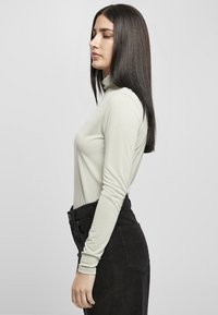Langærmet polo i lysegrøn stof, tætsiddende design. Kombineret med højtaljede sorte fløjl bukser, set fra siden, glat tekstur.