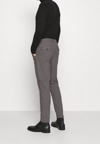 Person som bär grå slim-fit byxor med knappade bakfickor, svart turtleneck och svarta ankle boots, står mot en enkel vit bakgrund.