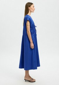 Blaues knielanges Kleid mit lockerer Passform, kurzen Ärmeln, gerafftem Bund mit einem Bindegürtel und glattem Stoff. Getragen mit schwarzen Flip-Flops.