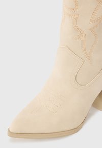 Bottine en daim beige avec un bout pointu et un talon bloc. Présente des coutures décoratives en motif ondulé sur le dessus.