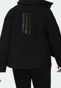 Personne portant une veste noire avec trois rayures réfléchissantes verticales sombres dans le haut du dos, se tenant contre un fond uni.