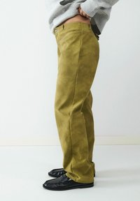 Oliwkowe spodnie wykonane z miękkiego, teksturowanego materiału; mają wysoki stan i kieszeń boczną; noszone z czarnymi butami typu slip-on.