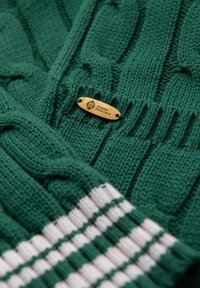 Suéter verde de punto con un diseño de cable texturizado, puños acanalados y una etiqueta de madera grabada con "Ombre Heritage". Detalles en rayas blancas.