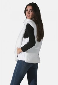 Gilet en fausse fourrure blanche avec un intérieur matelassé, doté d'un col montant et d'un design sans manches, associé à un haut à manches longues noir et un jean bleu.