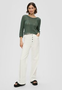 Grüner Strickpullover mit offener Maschenstruktur und Dreiviertelärmeln, kombiniert mit hoch taillierten weißen weit geschnittenen Hosen mit Knöpfen. Schwarze Sandalen.