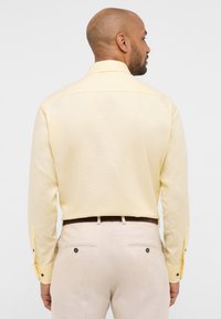 Camicia gialla chiaro a maniche lunghe con tessuto strutturato, polsini abbottonati e colletto standard, abbinata a pantaloni chiari con tasche posteriori.