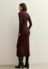 Abito lungo attillato in maglia bordeaux con maniche lunghe, scollatura alta e spacco laterale, abbinato a stivali neri lucidi con tacco al ginocchio.